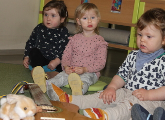 Drei kleine Kinder sitzen neben einer Gitarre, auf der ein Hase (Kuscheltier) spielt