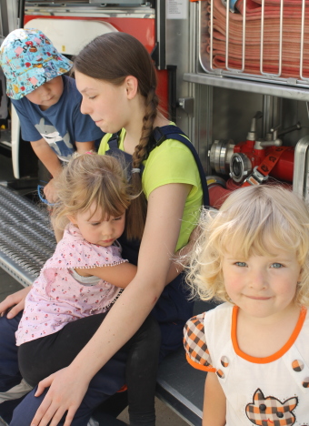 Auf dem Bild sind drei Kinder an einem Feuerwehrauto sitzen zu sehen