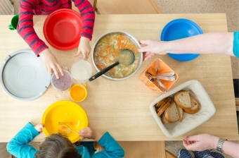 Zwei Kinder sind von oben zu sehen, sie sitzen an einem gedeckten Tisch mit einer Schüssel Suppe und mehreren Kannen mit Getränken vor sich auf dem Tisch