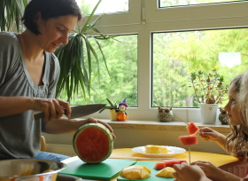 Auf dem Bild sind eine Frau und ein Kind zu sehen, die an einem Tisch Obst schneiden.